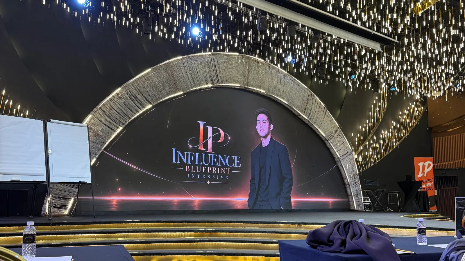 The stage at Confetti KL before a session of Jeff Leong's Founder IP course: an arch of lights, a screen with the course logo and his portrait, a ceiling of hanging lights.